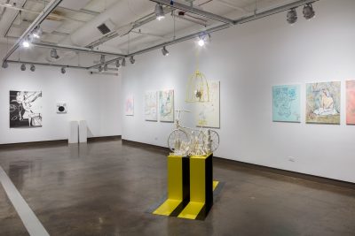 Installation view of various paintings in background. There is a black and yellow sculpture installed in the foreground with a small bicycle on top.