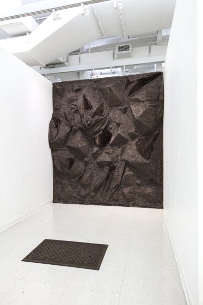A small brown rug sits at the front of a white room in which the far back wall has been transformed into a bulging rocky like terrain covered soil