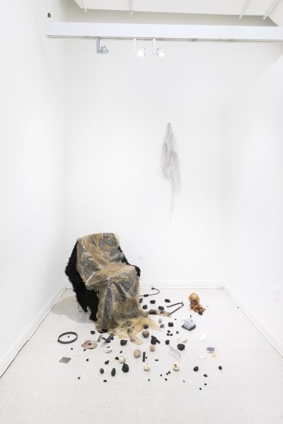 Keika OkamotoFabric and skin like material draped over a chair in the corner of a white room, with tiny found objects scattered on the floor