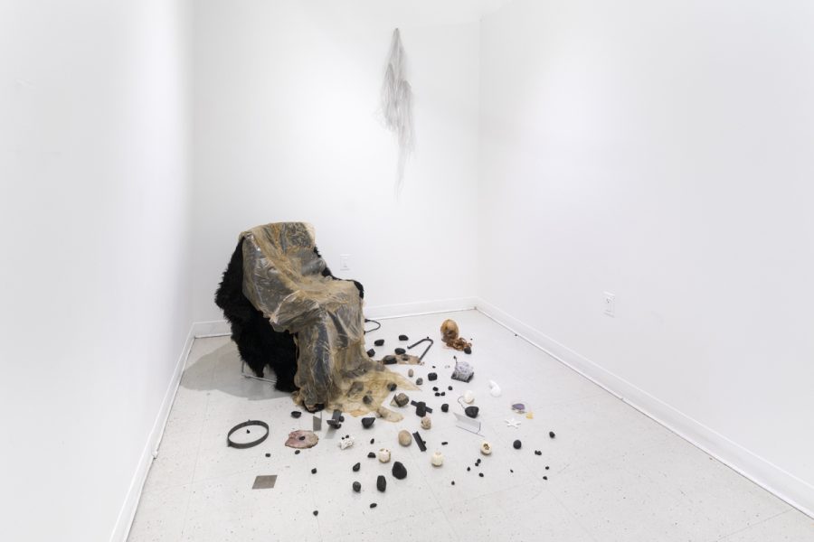 Fabric and skin like material draped over a chair in the corner of a white room, with tiny found objects scattered on the floor