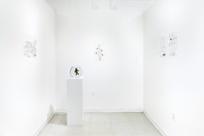 3 vacuum sealed sculptures holding water hang on the walls of a white room, with a pedestal in the center holding a man made out of algae floating in a fish bowl