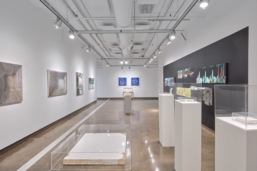 An installation view of an exhibition at the SVA Chelsea Gallery for the exhibition titled "Liminality". The view shows a white room with artworks hanging on the walls. In the foreground are three white pedastals with artwork on them and a plexiglass vitrine on the ground covering an artwork.