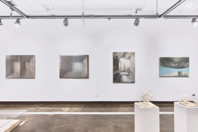 Four paintings depicting metallic and transparent surfaces installed on a white wall, with two pedestals displaying sculptures of a house
