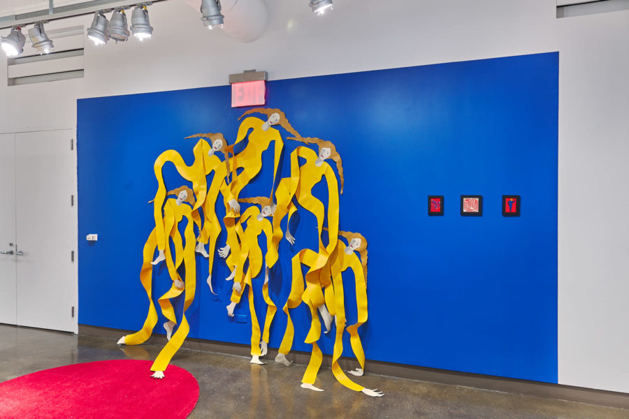 Exhibition Sticks and Stones May Break My Bones But Words Will never Hurt Me 2019. SVA Chelsea Galleries,New York. Installation view. Artwork by Annie (Flint) Kirschenbaum. Four female figures cut out of a plastic material partially mounted to a blue wall. Figures wearing a yellow garment.