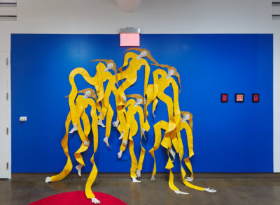 Exhibition Sticks and Stones May Break My Bones But Words Will never Hurt Me 2019. SVA Chelsea Galleries,New York. Installation view. Artwork by Annie (Flint) Kirschenbaum. Four female figures cut out of a plastic material partially mounted to a blue wall. Figures wearing a yellow garment.