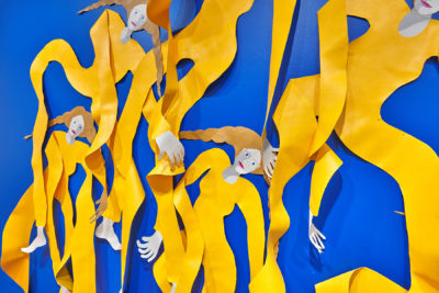 Exhibition Sticks and Stones May Break My Bones But Words Will never Hurt Me 2019. SVA Chelsea Galleries,New York. Installation view. Artwork by Annie (Flint) Kirschenbaum. Detail. Four female figures cut out of a plastic material partially mounted to a blue wall. Figures wearing a yellow garment.