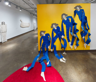 Exhibition Sticks and Stones May Break My Bones But Words Will never Hurt Me 2019. SVA Chelsea Galleries,New York. Installation view. Artwork by Annie (Flint) Kirschenbaum. Four female figures cut out of a plastic material partially mounted to a yellow wall. Figures wearing a blue garment.