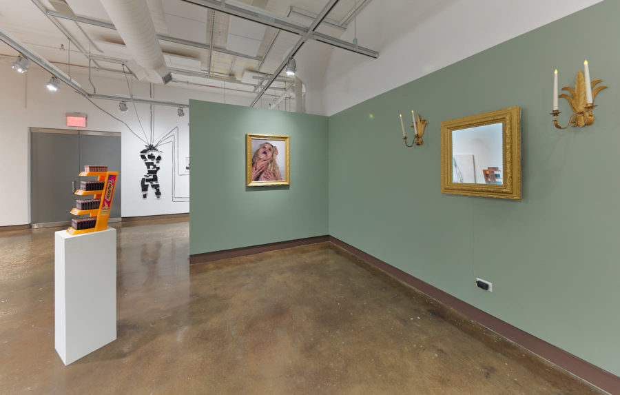 An installation view of an exhibition at the SVA Chelsea gallery titled "Sticks and Stones May Break My Bones, But Words Will Never Hurt Me". The view features a wall on the right hand side painted in a light green golor. On the right side a sculpture sits on a white pedestal. The artwork photographed in the center is by Hayley McCormack.