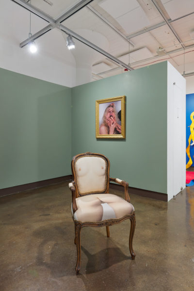 Exhibition Sticks and Stones May Break My Bones But Words Will never Hurt Me 2019. SVA Chelsea Galleries,New York. Installation view. Artwork by Hayley McCormack. A wooden chair with a nude woman torso printed on the fabric cushion. Digital photographic print of a portrait of a woman with her mouth open and hands grabbing her mouth. Gold frame.