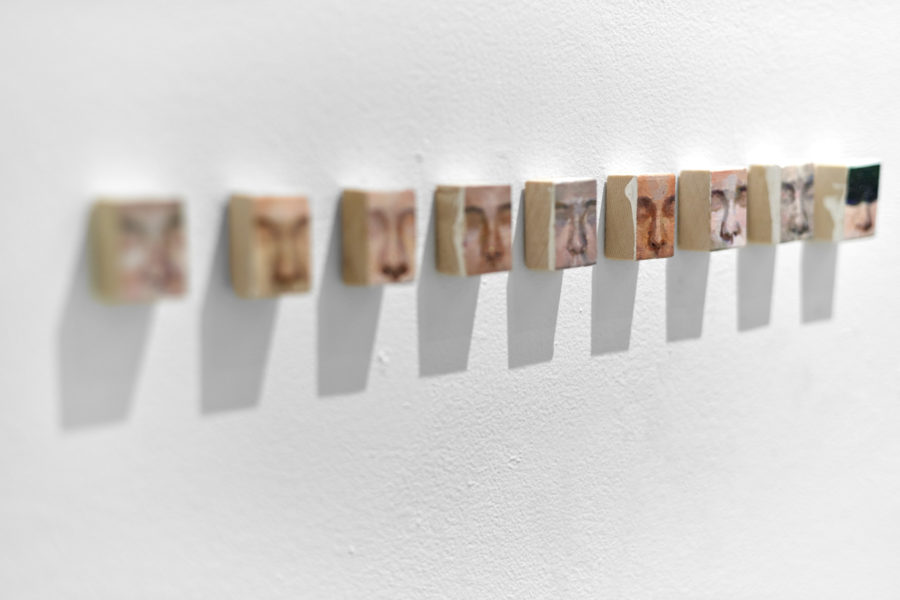 Artwork by Ruofan Chen. BFA Fine Arts, 2019. Nine close up portraits of a face on small pieces of wood mounted to a wall in a vertical column.