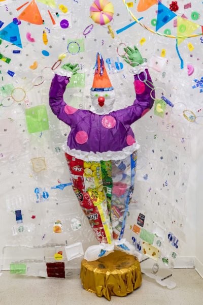 Artwork by Sophy Chang. BFA Fine Arts, 2019. Mixed media. Plastic pieces of material quilted into a large rectangular shape and a clown constructed out of colorful plastic.