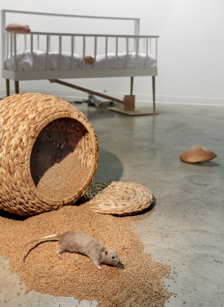 A sculpture resembling a foreshortened white bed in the background and an spilled basket of grain with a rat in the foreground.