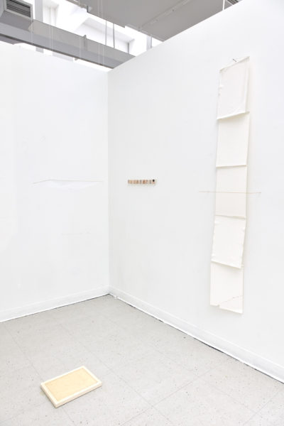 Artwork by Ruofan Chen. BFA Fine Arts, 2019. Installation view. Five sheets of paper placed on a vertical grid on a wall, white framed piece of yellow tinted paper on the floor and a small wooden object mounted to the wall.