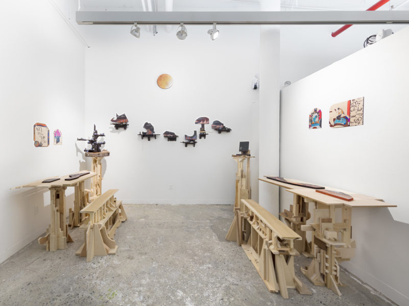 Installation view of multiple sculptures made out of wood that is painted, landscape imagery, multiple objects placed on the sculptures. Included are two assemblages of irregular pieces of lumber resembling a table and two benches.