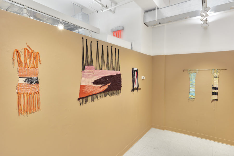 Installation view of multiple colorful textile artworks hanging on a flesh tone wall.