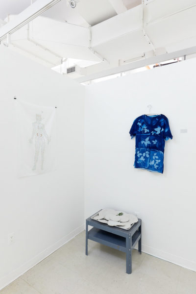 In the corner of a white room sits a small grey table with cement discs, above hangs a dark blue t-shirt with a bleached leaf pattern