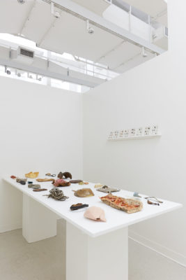 In a white room, a wall mounted shelf and table hold sculptures made of found natural materials