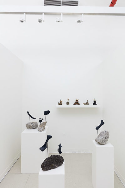 In a white room, a wall mounted shelf and 3 pedestals hold sculptures made of found natural materials
