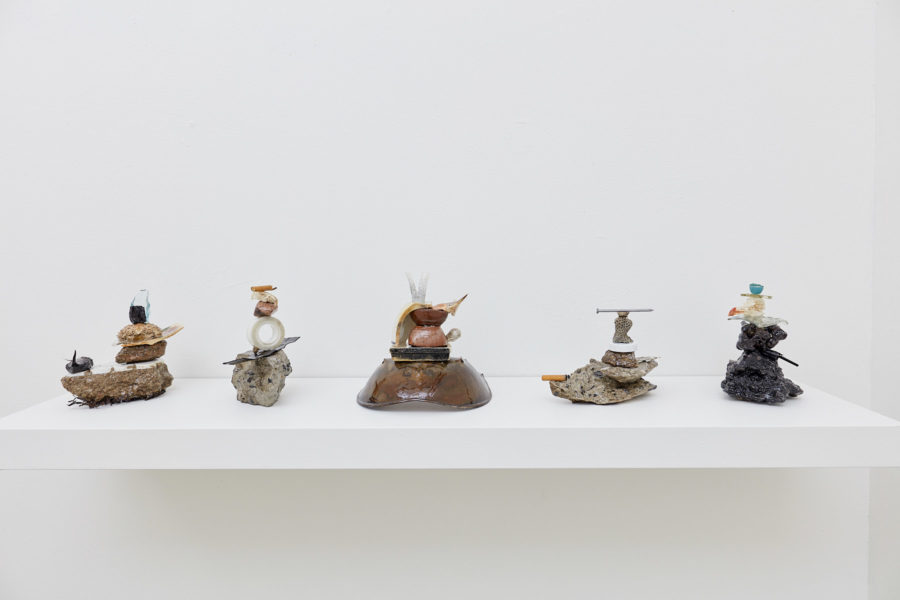 Five sculptures rest on a shelf. Each sculpture is composed of neatly stacked found objects, coated in resin.