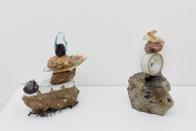 Two sculptures composed of neatly stacked found objects, such as broken china, bird skeleton, sea weed and scotch tape