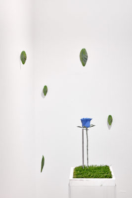 3d printed leaves hang from fishing line in a white room next to a pedestal covered in astroturf with a blue rose made of steel.