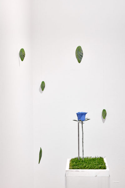 3d printed leaves hang from fishing line in a white room next to a pedestal covered in astroturf with a blue rose made of steel.