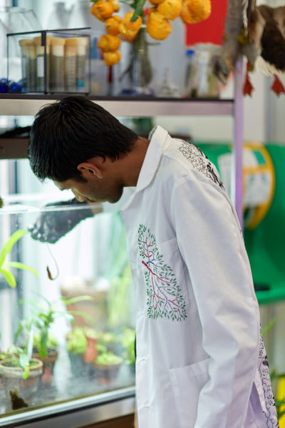A man in an embroidered lab coat stands in front of a window with his hands in his pockets