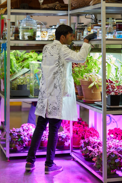 A man in an embroidered lab coat stands in the corner of brightly lit shelves filled with plants and specimens