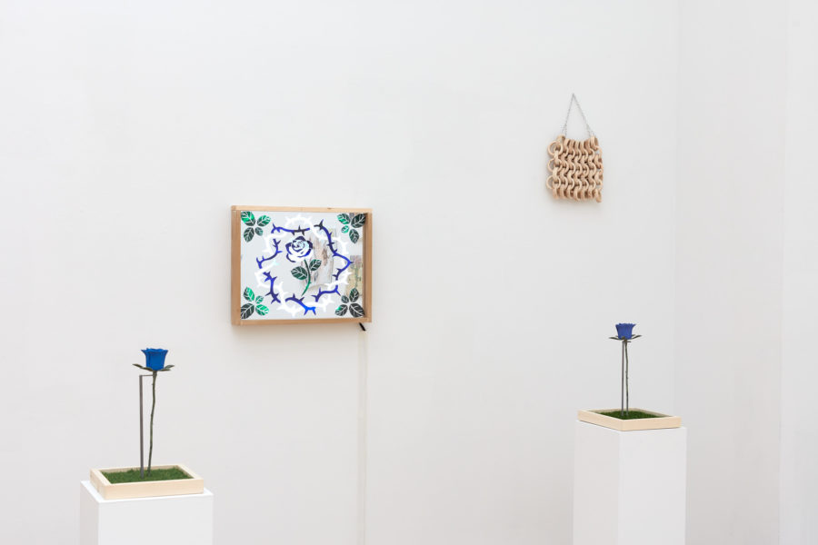 Exhibition Tales and Whispers 2019. SVA Flatiron Gallery, New York. Artwork by Emma Fasciolo. A framed mixed media artwork with an abstract blue flower and barbed wire, Two blue flower sculptures on pedestals and ceramic made chain-male mounted on the wall. .