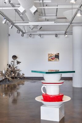 A sculpture that depicts large bowls and cups stacked on top of each other. At the top is a table. The sculpture is in an open gallery. Two artworks are seen in the background of the photograph.