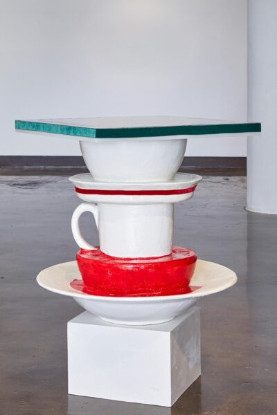A sculpture that depicts large bowls and cups stacked on top of each other. At the top is a table. The sculpture is in an open gallery.