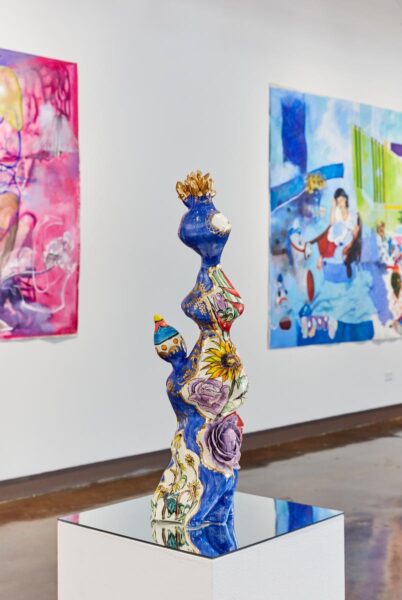 A colorful ceramic organic sculpture installed on a pedestal with a mirror and 2 large paintings in the background