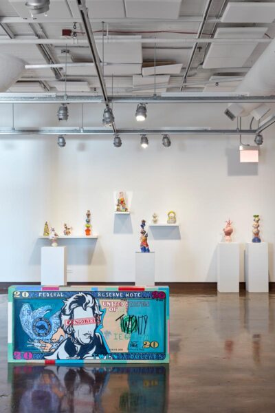Installation view with a large bill note sculpture on the floor and organic colorful ceramic sculptures in the background.