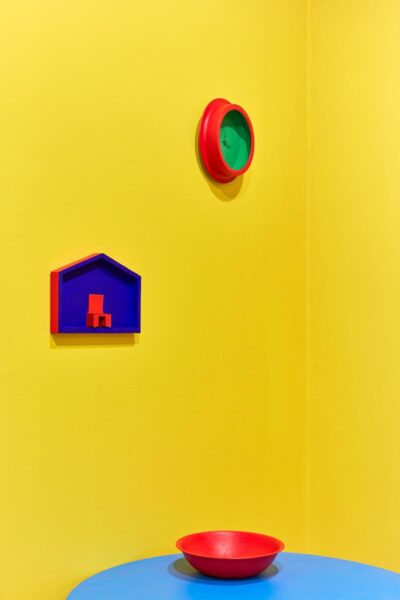 Installation view of a bright yellow corner with three green, red and blue painted sculptures.