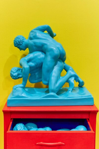 Replica of a classic greek sculpture of two naked men fighting painted in bright light blue over a saturated red chest of drawings.
