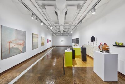 Installation view of paintings hung on a white wall on the left side of the frame, and in the middle are two green pedestals with miscellaneous objects on them and other things.