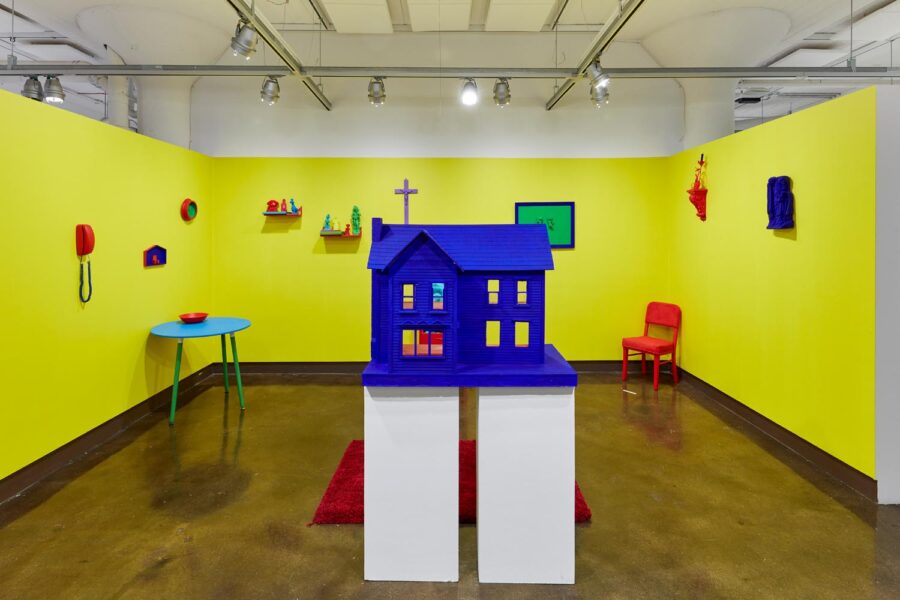 Exhibition view of "Chromaphilia/Chromophobia" of a yellow rooms with many small home objects on the walls and floor rendered in primary colors. In the foreground is a primary blue dollhouse.