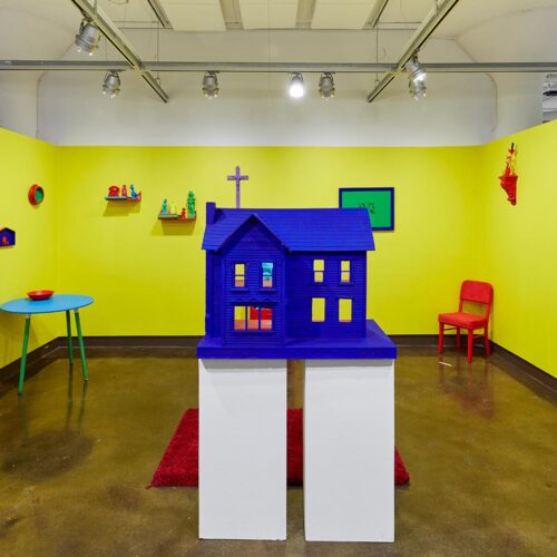 Exhibition view of "Chromaphilia/Chromophobia" of a yellow rooms with many small home objects on the walls and floor rendered in primary colors. In the foreground is a primary blue dollhouse.