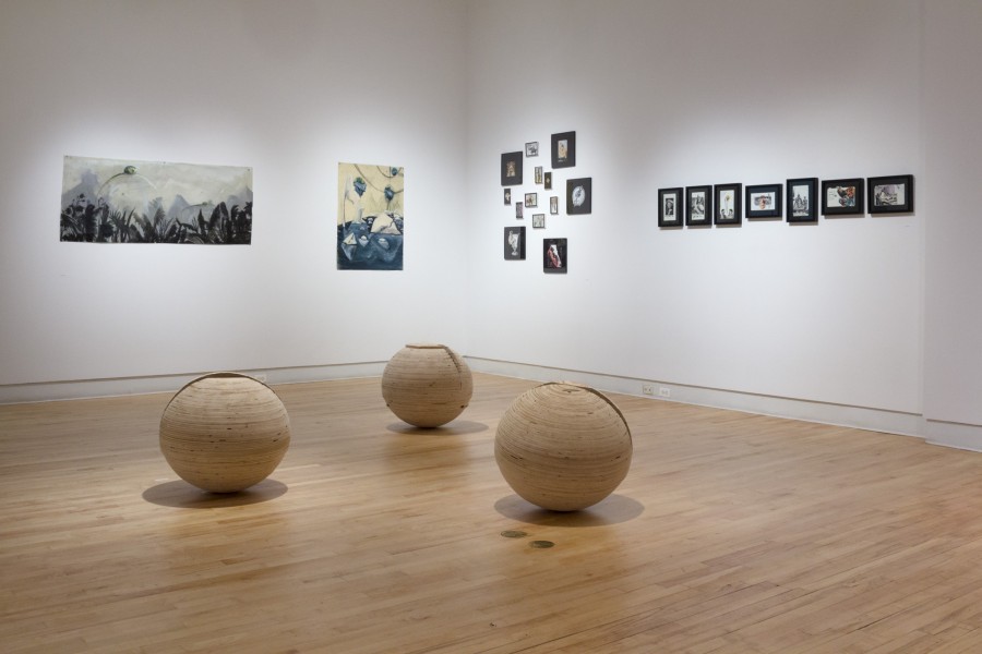 Installation view of three large wooden balls on the floor and multiple paintings and print hung on the background wall.