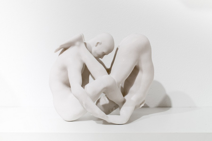 Anthropomorphic sculpture of two people with hands glued together and one person sitting on the ground with head tilted forward, and the other person is standing with feet on the shoulders of first person and the head on the floor.
