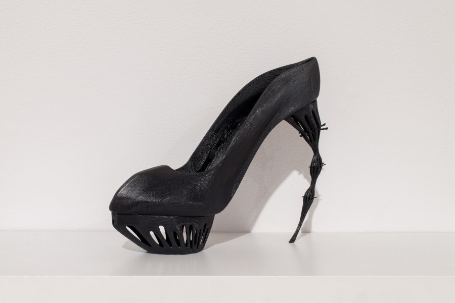 A black platform heeled shoe with the head made thick at the base of the shoe, and it is getting thinner at the end of it where it sits on the floor.