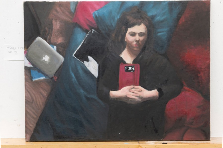 A woman painted while is laid on her back on the bed with blue and red sheets and pillows. She holds her phone in a red case with both hands on her chest. She wears a black shirt. A MacBook on the floor and some notebooks under it are near the bed.