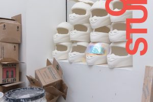 Open Studios poster with an illustration containing ceramic sculptures of motocross helmets stacked near a window and cardboard boxes