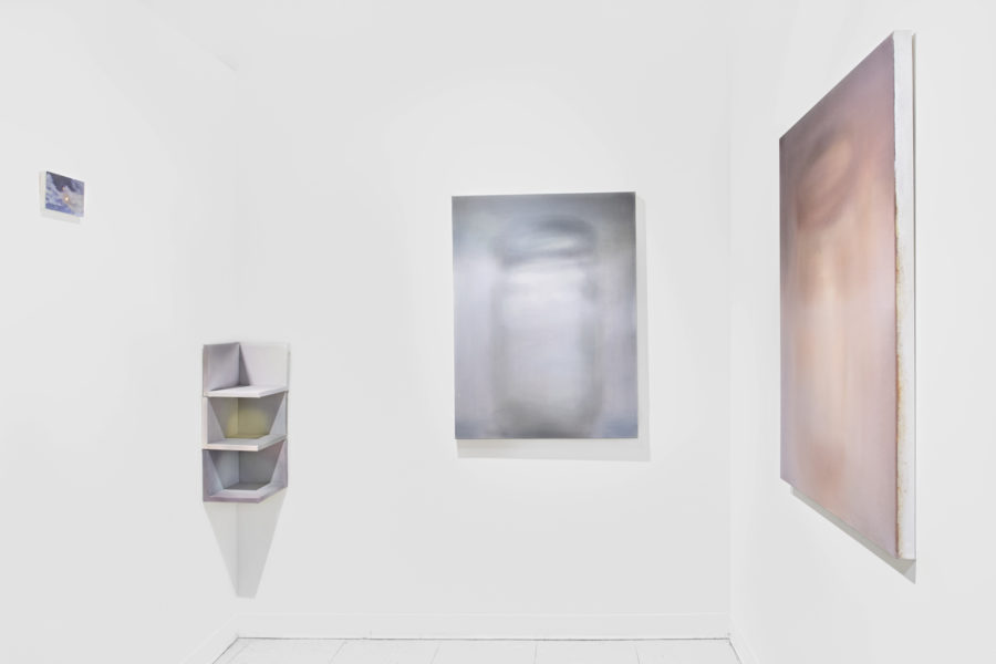 An installation view showing three abstract paintings and one three-tier corner shelf installed in a white-walled studio.