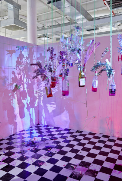 Artwork by Ron Burgos Cavalié. BFA Fine Arts, 2019. Installation view. Multiple lights of different colors, black and white checkered floor, projection of a face, and glass bottles with flowers in them hanging from the ceiling.