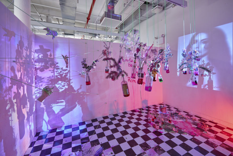 Artwork by Ron Burgos Cavalié. BFA Fine Arts, 2019. Installation view. Multiple lights of different colors, black and white checkered floor, projection of a face, and glass bottles with flowers in them hanging from the ceiling.