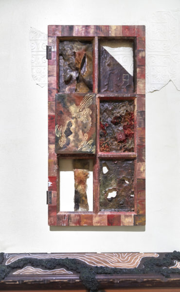 Wall mounted sculpture on wall that appears to be a door with inner squares containing amorphous sculptures that exist within the frame and on the wall behind the sculpture.