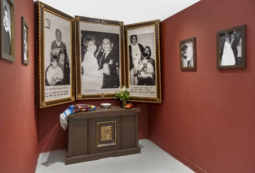 Installation of black and white wedding / family portraits framed and hung on adjacent walls. A triptych of large black and white photographs are opened above a dresser which also holds a folded Mexican blanket, a ceramic bowl, and a vase of flowers.