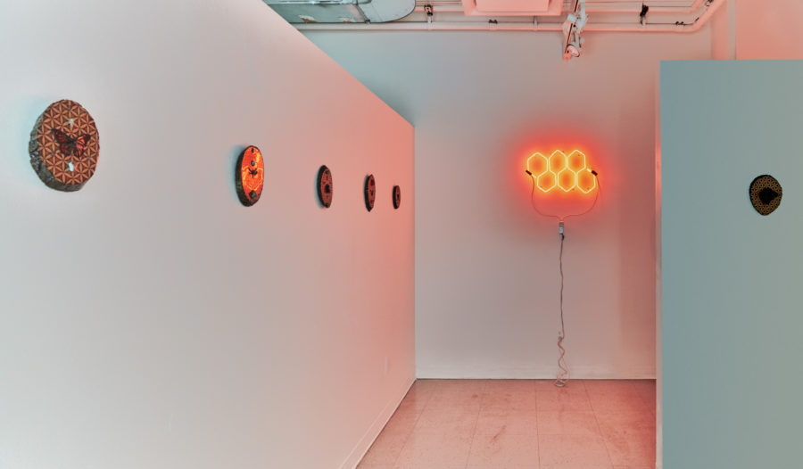 Artwork by Melissa Rose Pressler. BFA Fine Arts, 2019. Installation view. Multiple circular mixed media artworks hanging on the wall. Neon sign with six orange hexagons.