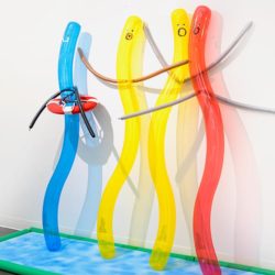 Sculpture by Zidong Chen. The sculpture is made of mixed materials. In the center are, what look like, four long and narrow ballow shapes with a face drawn on the ends with a black marker. The forms are shaped in curvy waves and have long thin attachments to them that allude to arms. The shapes are blue, yellow, and red.